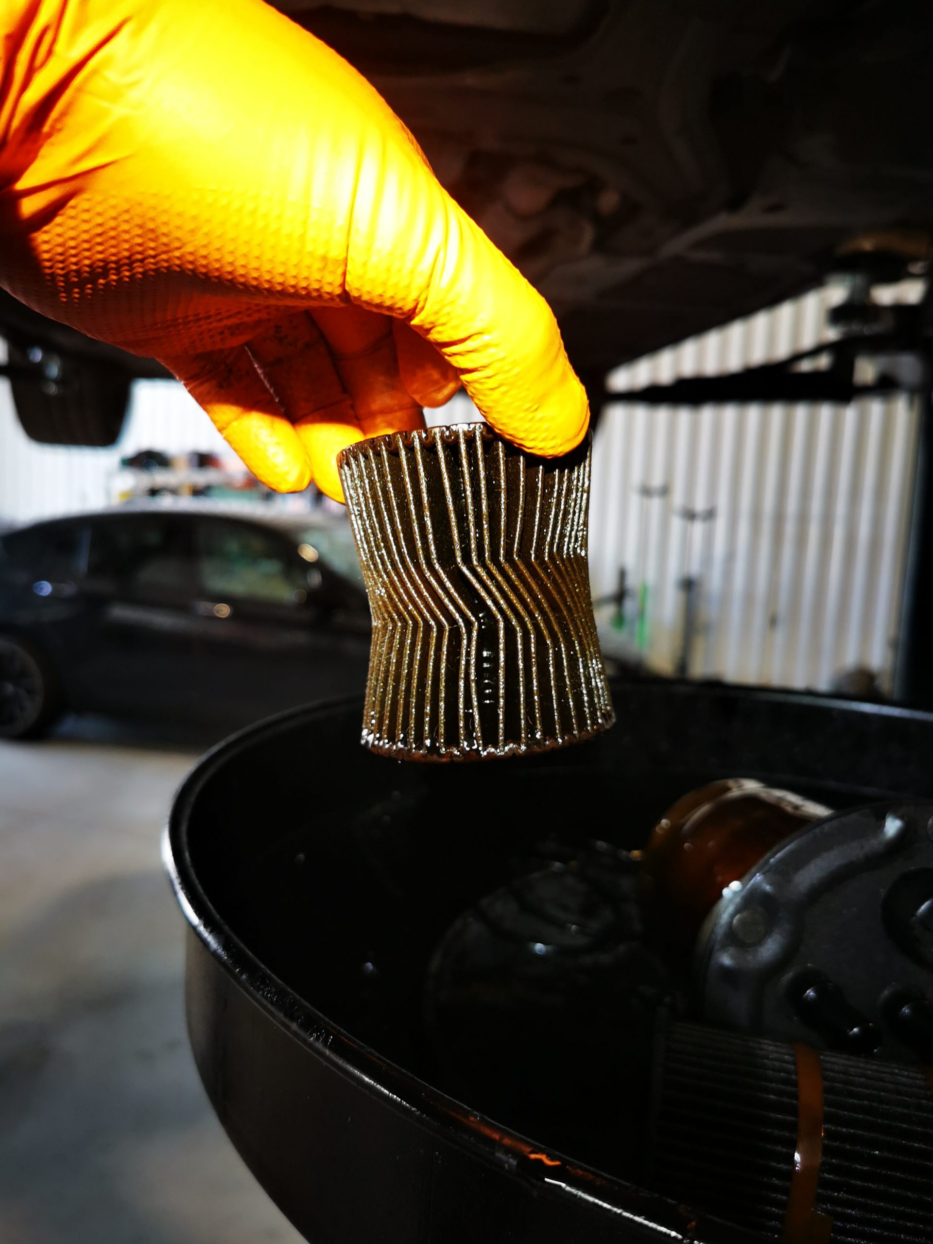 Gloved hand holding a used pleated oil filter element covered in oil above an oil drain pan during an engine oil change in a garage